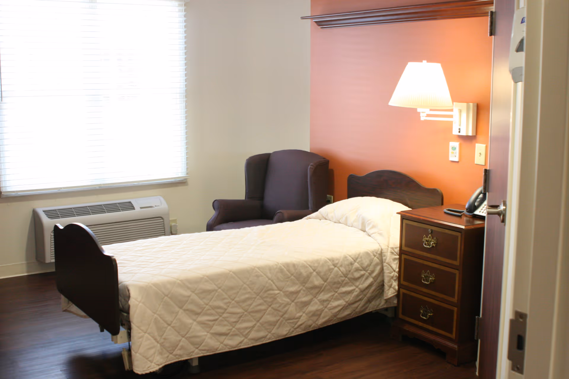 A simple bedroom in a senior living facility with a single bed covered in a white quilt, a dark wooden nightstand with three drawers, a wall-mounted lamp, a dark upholstered armchair, and a window with blinds letting in natural light.