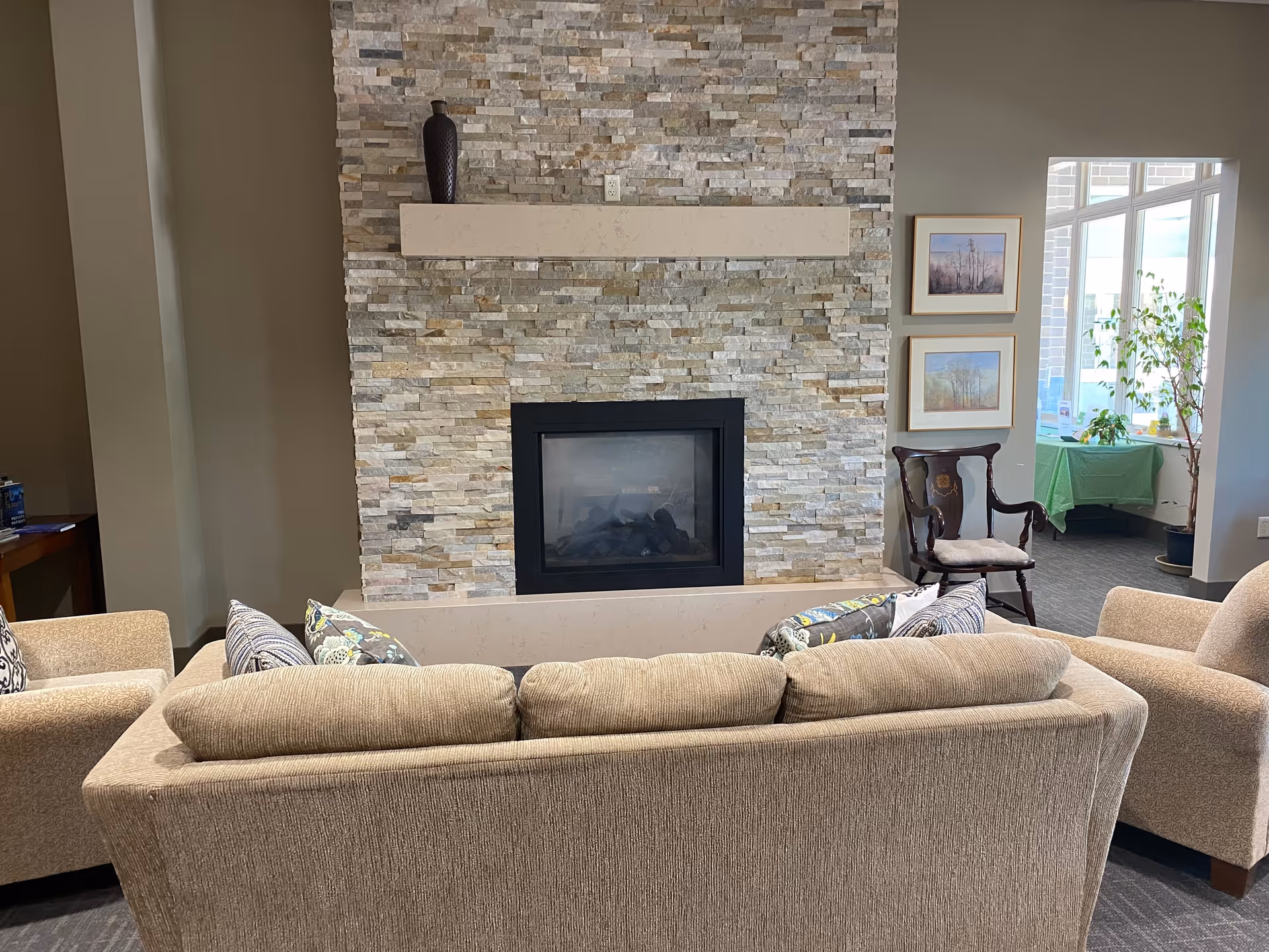 A cozy living room area featuring a beige sofa with patterned cushions facing a stone fireplace with a black frame. To the right, there is a wooden chair with a cushion, two framed pictures on the wall, and a window letting in natural light. A small table with a green tablecloth and plants is visible in the background.