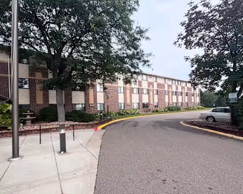 Exterior view of a brick senior living facility with a curved driveway, trees, and a sidewalk.