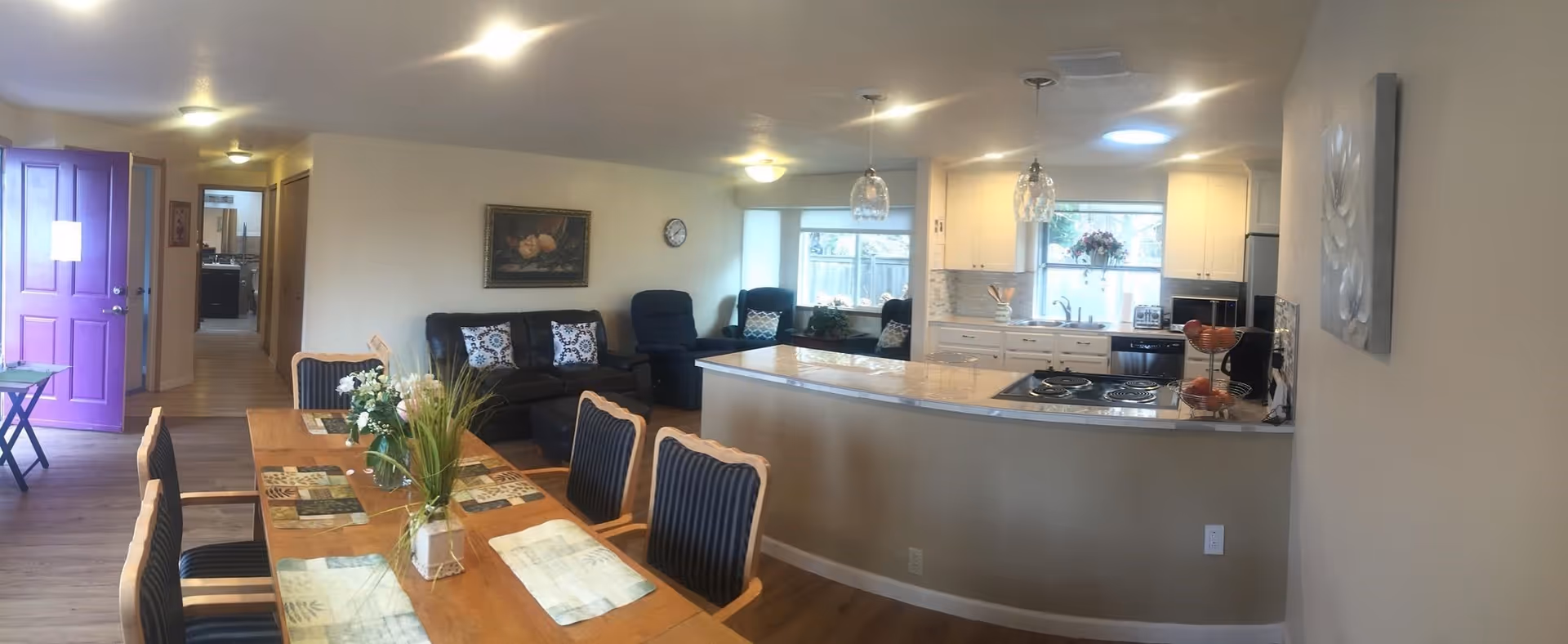 Interior view of a senior living facility showing a combined dining and living area with a kitchen. The dining area has a wooden table with six chairs and floral centerpieces. The living area has a black leather sofa and two recliners near a window. The kitchen features white cabinets, a countertop with a built-in stove, and hanging pendant lights. A purple door is open, revealing a hallway with additional rooms.