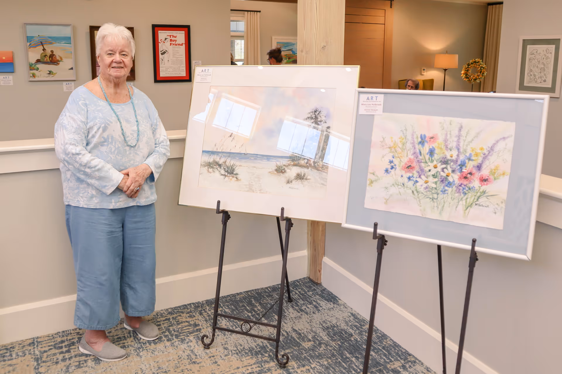 An elderly woman stands indoors next to two easels displaying framed watercolor paintings, one of a beach scene and the other of a colorful bouquet of flowers. The room has light-colored walls with additional artwork hanging and a carpeted floor.