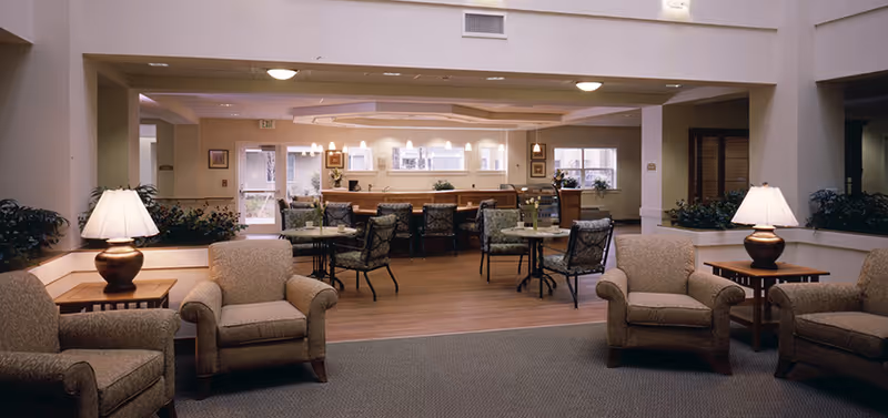 Open common area with upholstered armchairs in the foreground, small dining tables and chairs, and a serving counter/bar in the background.