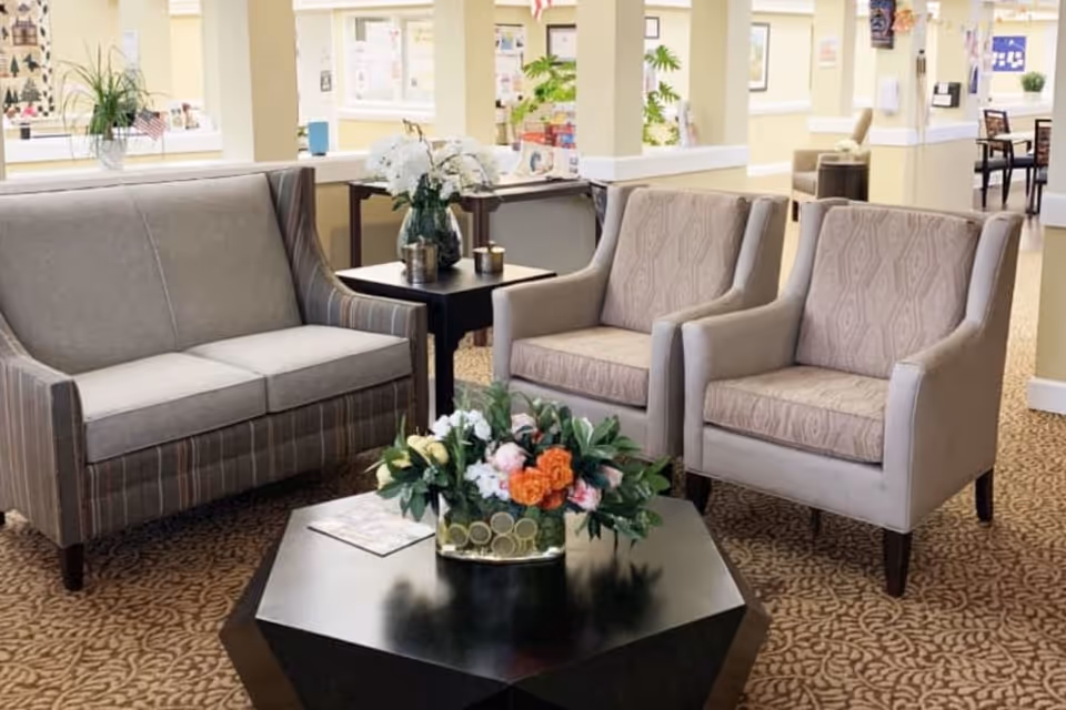 A seating area in a senior living lobby with a sofa, two armchairs, a central coffee table holding a floral arrangement, and patterned carpet.