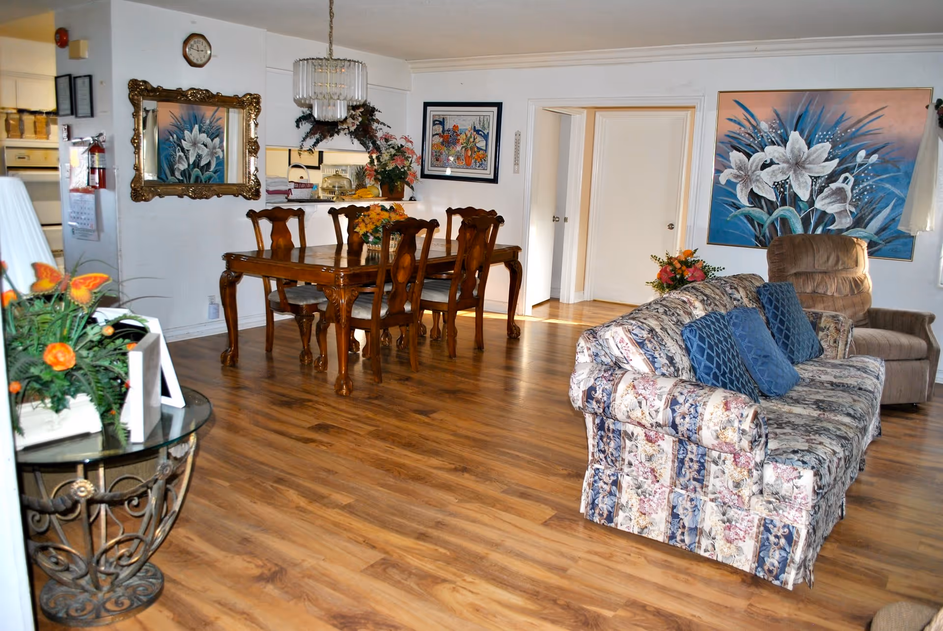 A cozy living and dining area with wooden flooring. The dining area features a wooden table with six chairs and a floral centerpiece. The living area has a floral-patterned couch with blue cushions and a brown recliner chair. The walls are decorated with large floral paintings and a mirror with a gold frame. There is a glass side table with a flower arrangement and picture frames. The kitchen is partially visible through a pass-through window behind the dining table.
