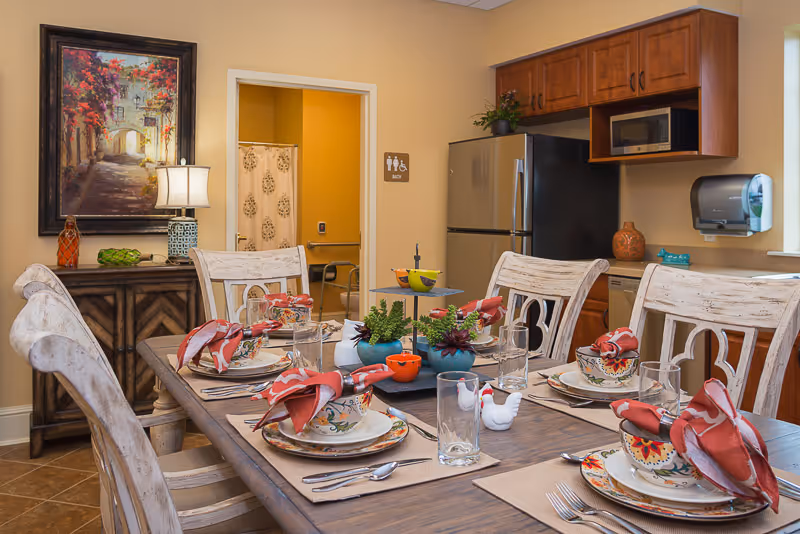 A dining area in a senior living facility with a wooden table set for six people. Each place setting includes a colorful bowl, plate, glass, silverware, and a red cloth napkin. The table is decorated with small potted plants and ceramic figurines. In the background, there is a kitchen area with wooden cabinets, a stainless steel refrigerator, a microwave, and a paper towel dispenser. A doorway leads to a bathroom with a shower curtain visible. A framed painting and a lamp are on a sideboard against the wall.