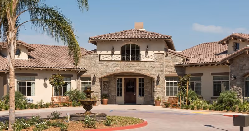 Front exterior view of a senior living facility with a stone and stucco facade, tiled roof, a central entrance with double doors, potted plants, benches, palm trees, and a circular driveway with a fountain in the center.