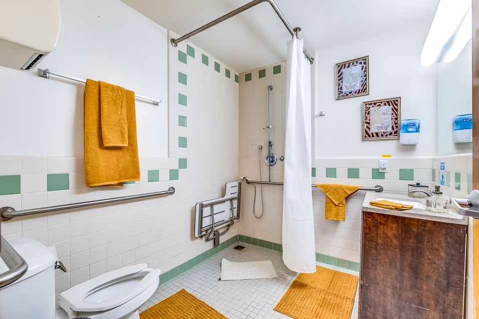 A senior-friendly bathroom with white and green tiled walls, a walk-in shower with a white curtain, grab bars, a fold-down shower seat, a toilet with grab bars, a wooden vanity with a sink, and mustard yellow towels and bath mats.