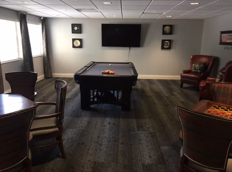 Interior common room with a pool table centered under a wall-mounted TV, surrounded by chairs and tables.