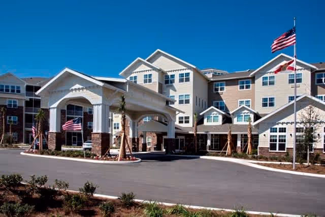 Exterior view of a multi-story senior living facility with a covered entrance, landscaped grounds, and American flags flying on flagpoles under a clear blue sky.