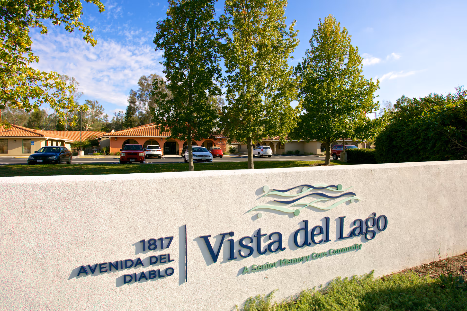 Exterior view of Vista del Lago Memory Care facility with a white sign displaying the name and address 1817 Avenida Del Diablo. The background shows a parking area with several cars and buildings with red-tiled roofs, surrounded by green trees and a partly cloudy sky.