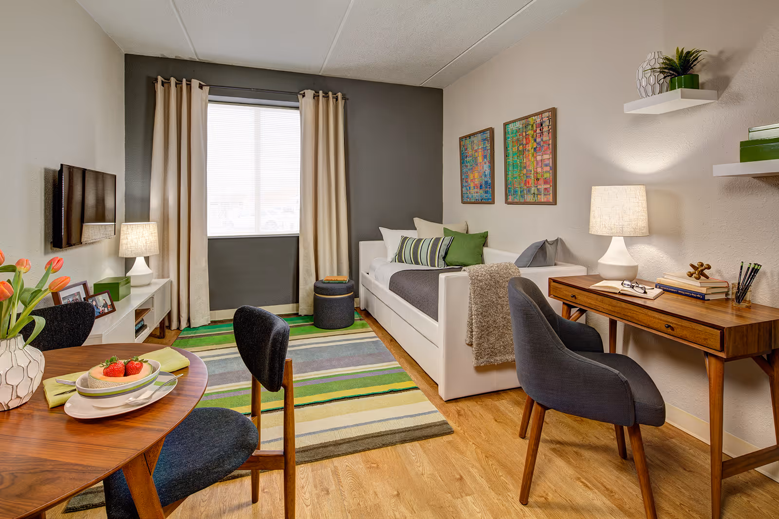 Bright furnished living room/studio with a daybed, desk, dining table, rug and wall-mounted TV.