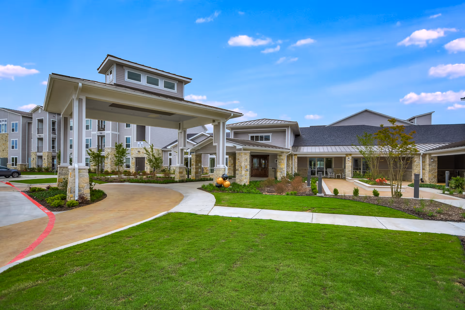Exterior view of Legacy Oaks of Azle Senior Living facility showing a covered entrance with stone pillars, a driveway, well-maintained green lawn, and a multi-story building under a blue sky with scattered clouds.