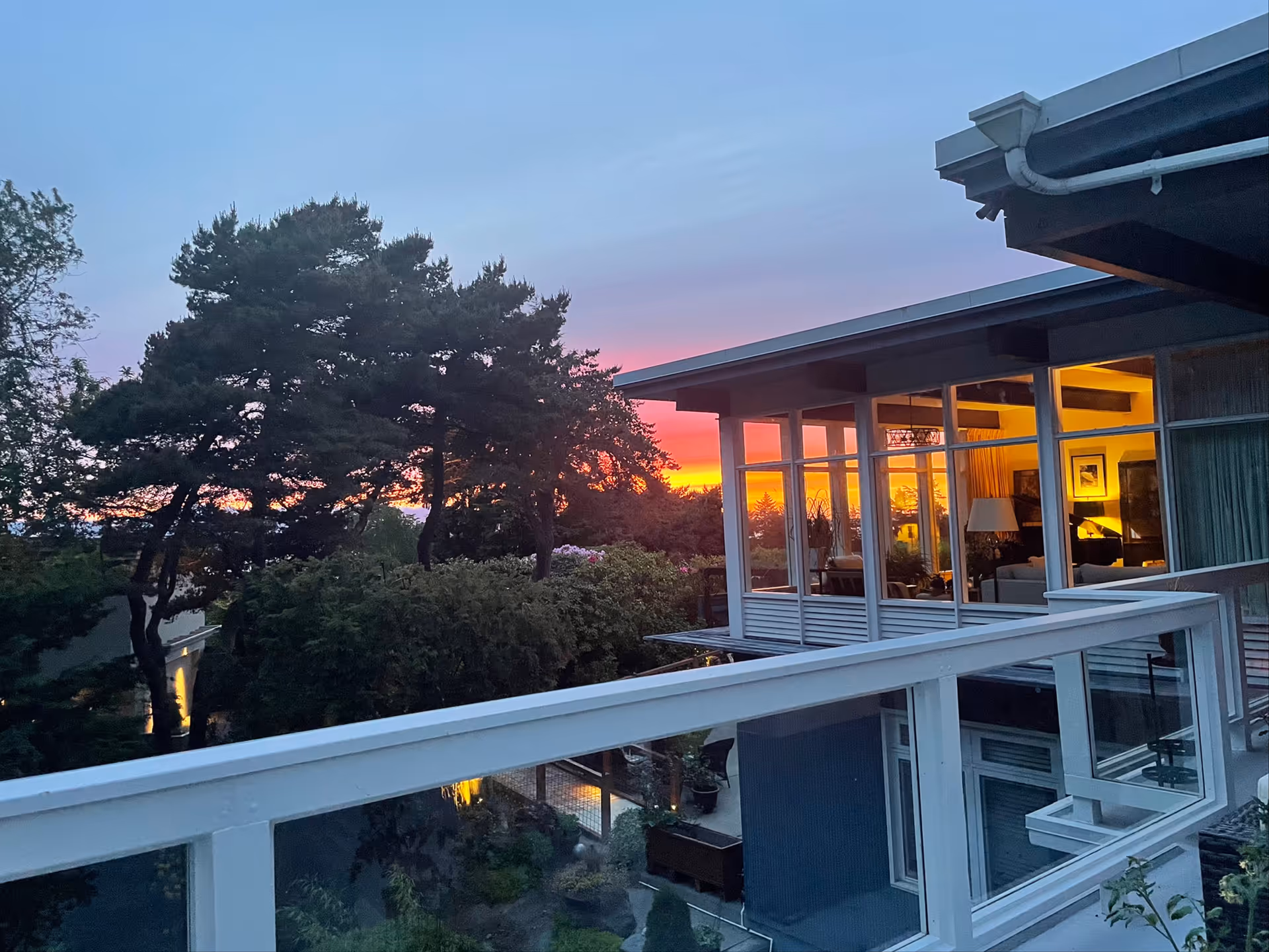 View from a balcony overlooking a garden with trees at sunset. The sky is colorful with shades of orange, pink, and purple. Part of a modern building with large windows is visible, showing a warmly lit interior with furniture and a lamp.