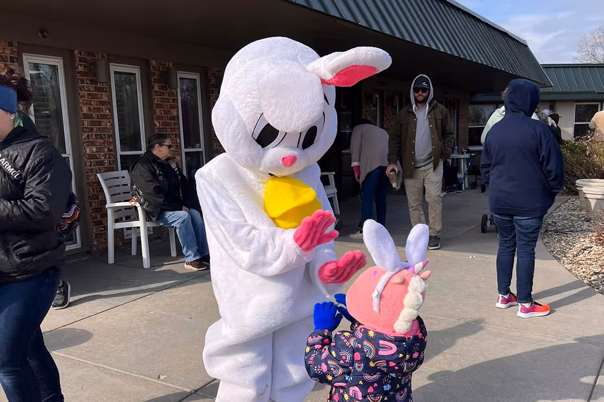 A person in a white bunny mascot interacts with a small child outside a brick building.