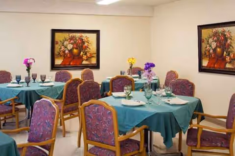 A dining room with teal tablecloths, purple chairs, vases of flowers, and framed paintings.