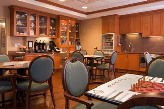 A cozy dining area with wooden cabinets and multiple round tables surrounded by green cushioned chairs. The room features a coffee station with coffee pots on a counter, a small refrigerator, a sink, and a microwave. One table in the foreground has a chessboard and some chess pieces set up.