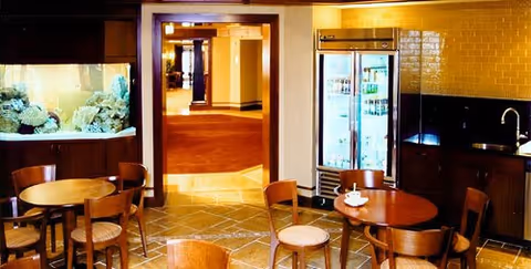Small dining/lounge area with round wooden tables and chairs, an aquarium, a refrigerated beverage cooler, and a sink/counter near a hallway.