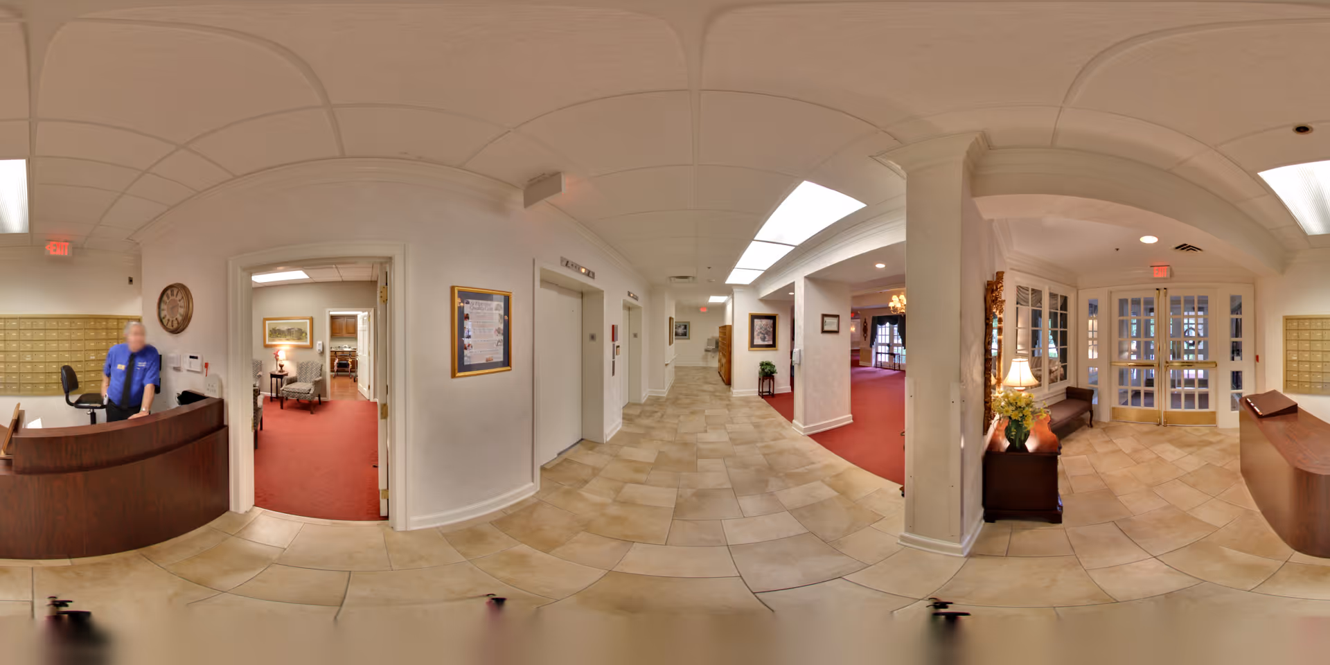 Panoramic view of the interior lobby area of The Lafayette senior living facility, showing a reception desk with a staff member, tiled flooring, white walls, elevators, seating areas with red carpet, framed artwork, and glass doors leading outside.