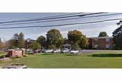 Exterior view of a senior living facility named Blocher Homes showing a parking lot with several cars, a green lawn in the foreground, and a two-story brick building with trees in the background under a clear sky.