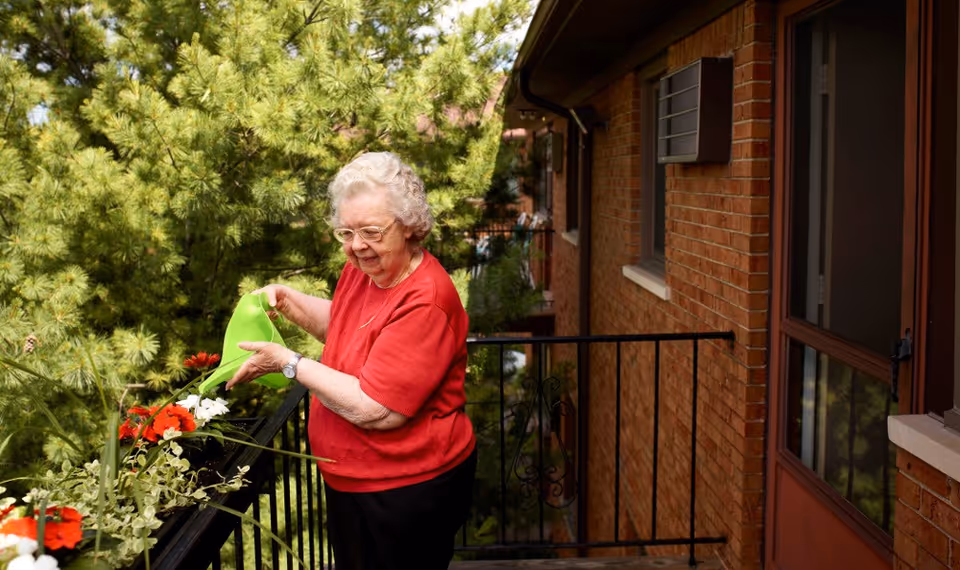 An elderly woman wearing a red shirt and glasses waters flowers on a balcony with a green watering can. The balcony has a black metal railing and is attached to a brick building. There are green trees in the background.