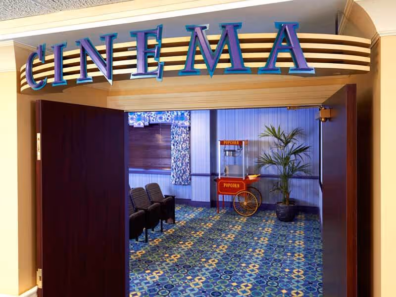 Entrance to a small cinema room with a sign above the door reading 'CINEMA' in large blue letters. Inside, there are a few rows of black theater seats, a popcorn machine on a red cart, a potted plant, and patterned blue carpet and curtains.