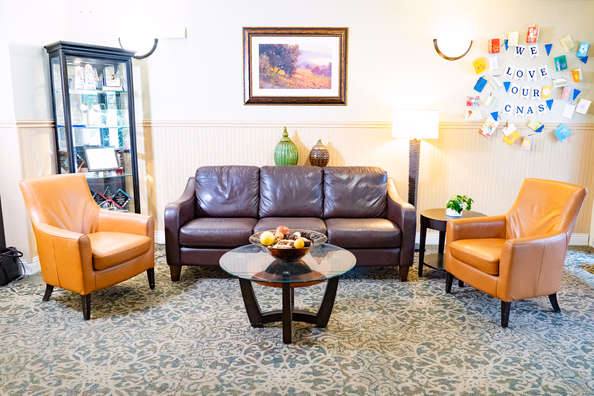 A cozy seating area in a senior living facility featuring a dark brown leather sofa flanked by two tan leather armchairs. In front of the sofa is a round glass coffee table with a decorative bowl of fruit and pinecones. Behind the sofa, there is a framed landscape painting on the wall and two decorative vases on a ledge. To the right, a floor lamp illuminates the space next to a small round side table with a potted plant. On the wall, colorful cards arranged in a circle spell out 'WE LOVE OUR CNAS'. A glass display cabinet with awards and memorabilia is visible on the left side of the image. The room has patterned carpet and light-colored walls with wainscoting.