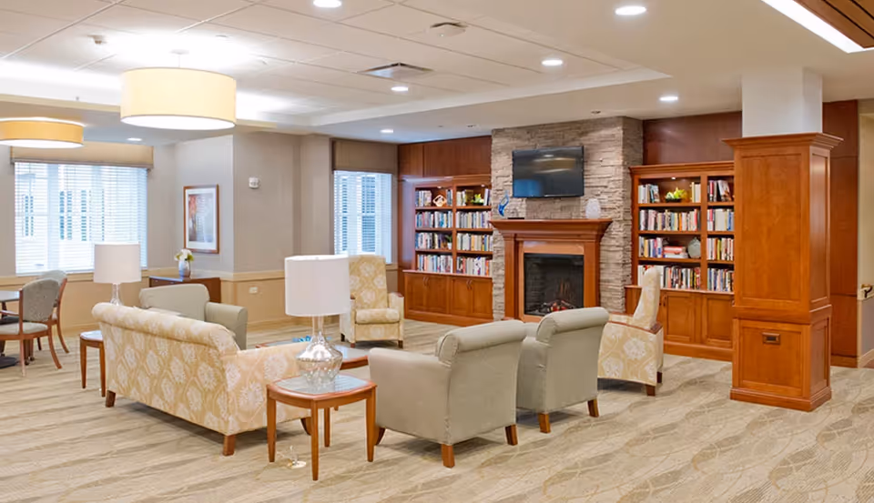 A spacious senior living common room with sofas and chairs arranged around a fireplace and built-in bookshelves.