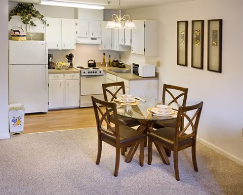 Open kitchenette with white cabinets and appliances beside a glass-top dining table set with four wooden chairs.
