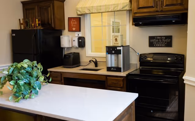 A kitchen area with dark wooden cabinets, a black refrigerator, a black stove, a countertop with a green leafy plant, a small sink, a coffee machine, and wall decorations including a sign that says 'I like my cream & sugar with a touch of COFFEE.'