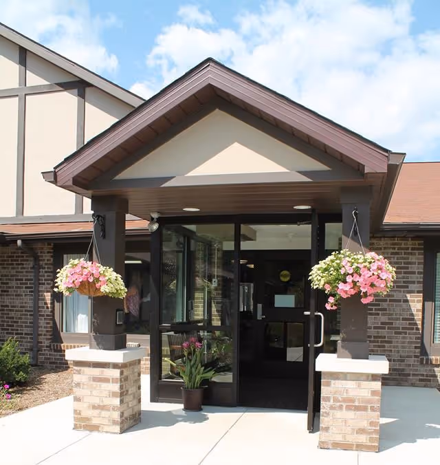 Entrance of a senior living facility with a covered porch supported by two brick pillars. Each pillar has a hanging basket with pink and white flowers. There is a potted plant on the ground near the glass doors. The building has a brick exterior and a sloped roof under a partly cloudy sky.