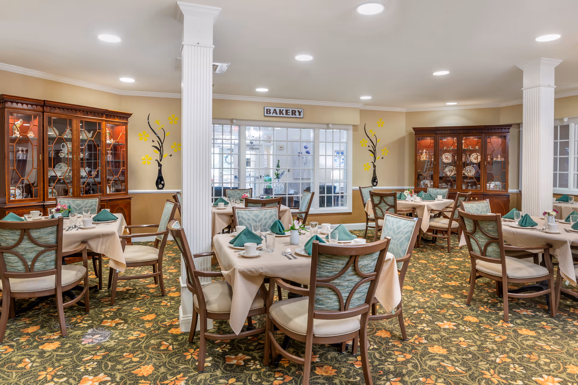 A dining room in a senior living facility with several tables covered in beige tablecloths, each set with green napkins, cups, glasses, and silverware. The room features floral patterned carpet, two large wooden china cabinets displaying dishes, and decorative wall art with yellow flowers and black vases. A sign above a windowed wall reads 'BAKERY'.
