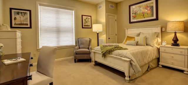 Well-lit furnished bedroom with a bed, nightstands, an armchair, a desk, and framed artwork on the walls.