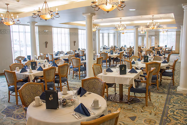 Spacious, well-lit dining room with multiple round tables set with white tablecloths, blue napkins, and wooden chairs.