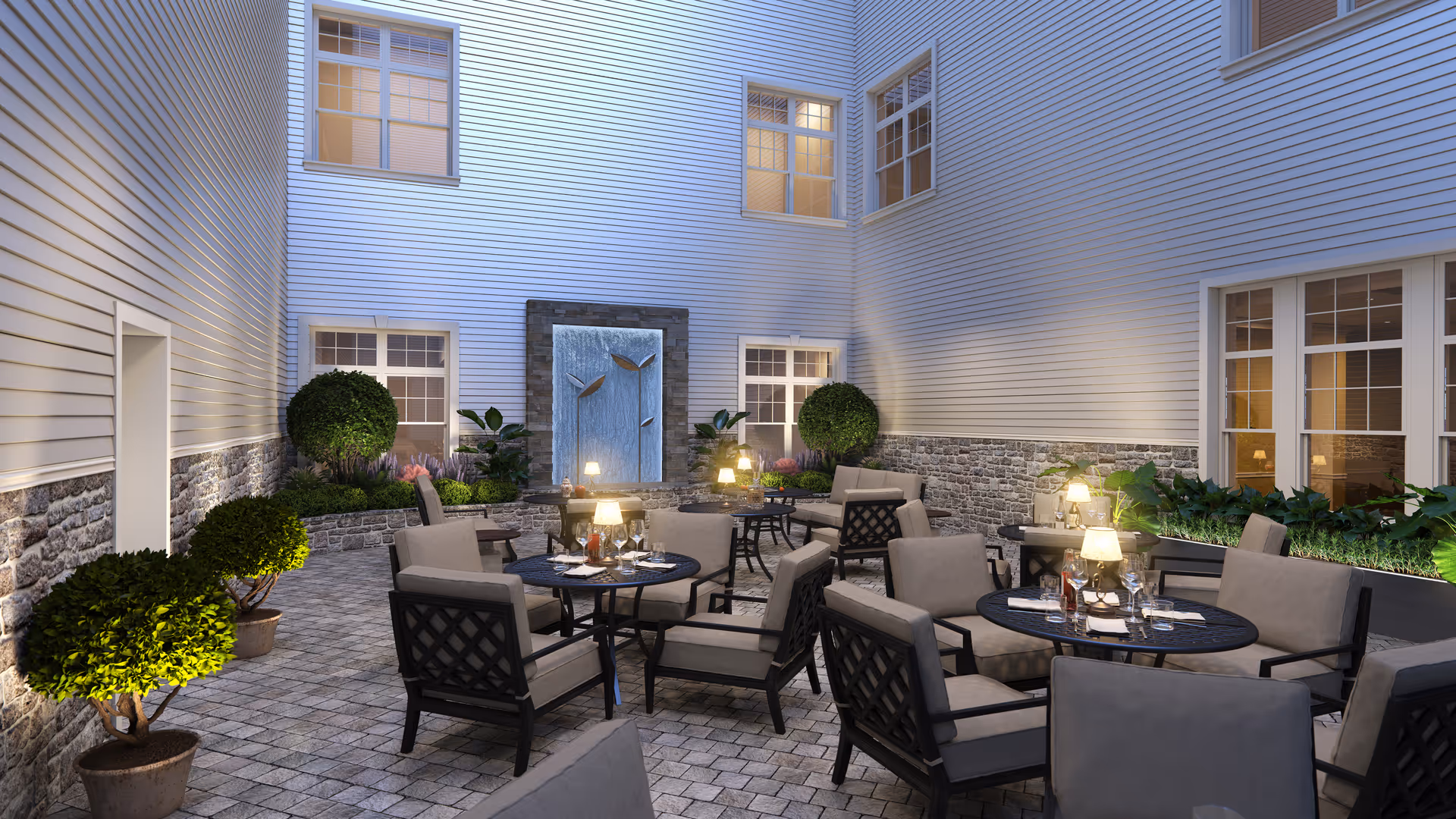 Outdoor courtyard area with several round tables and cushioned chairs arranged for seating. The space is surrounded by a building with multiple windows and features a decorative water fountain on one wall. There are potted plants and greenery along the edges, and small lamps on the tables providing warm lighting.
