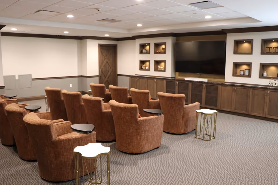 A cozy media room with two rows of brown upholstered armchairs facing a large flat-screen TV mounted on a wall with built-in shelves and cabinets. The room has a patterned carpet, recessed lighting, and a wooden door on the left side.