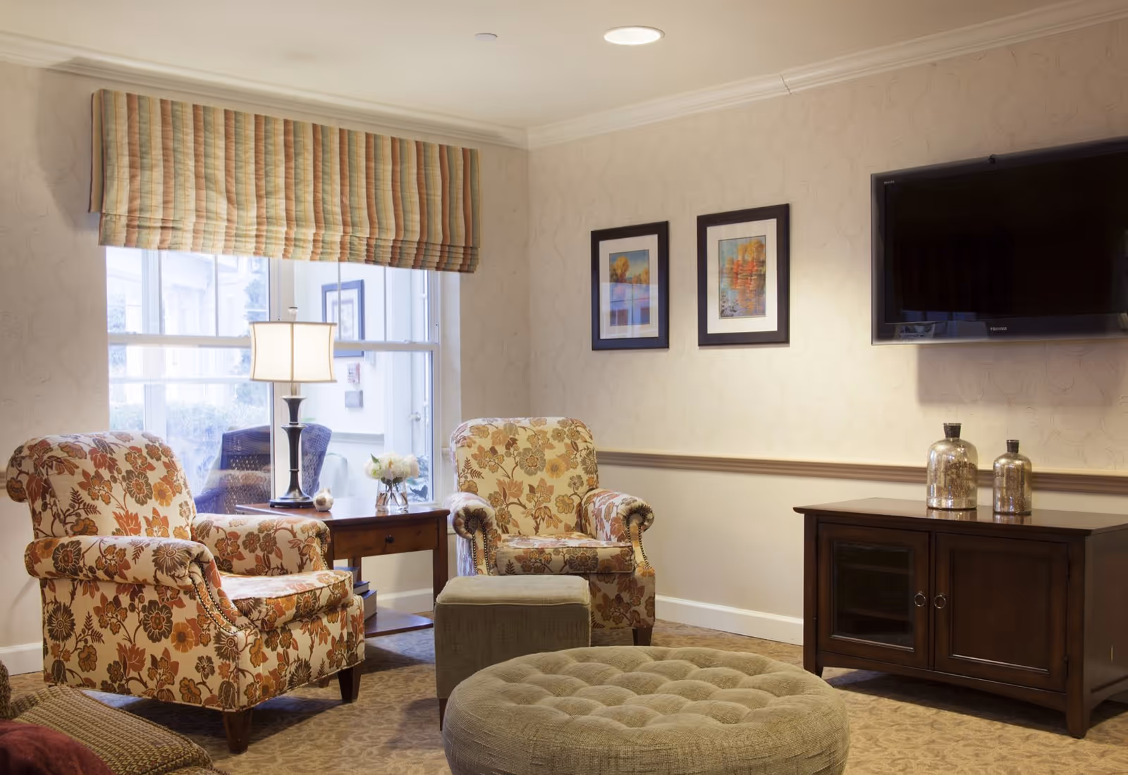 Cozy sitting area with two floral armchairs, a round tufted ottoman, side table with lamp, wall-mounted TV, and cabinet.