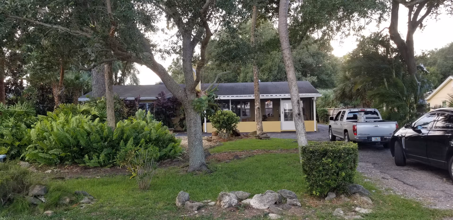 View of a single-story building with a screened porch surrounded by trees and greenery. There are two vehicles parked on a driveway to the right of the building, and a well-maintained lawn with bushes and rocks in the foreground.