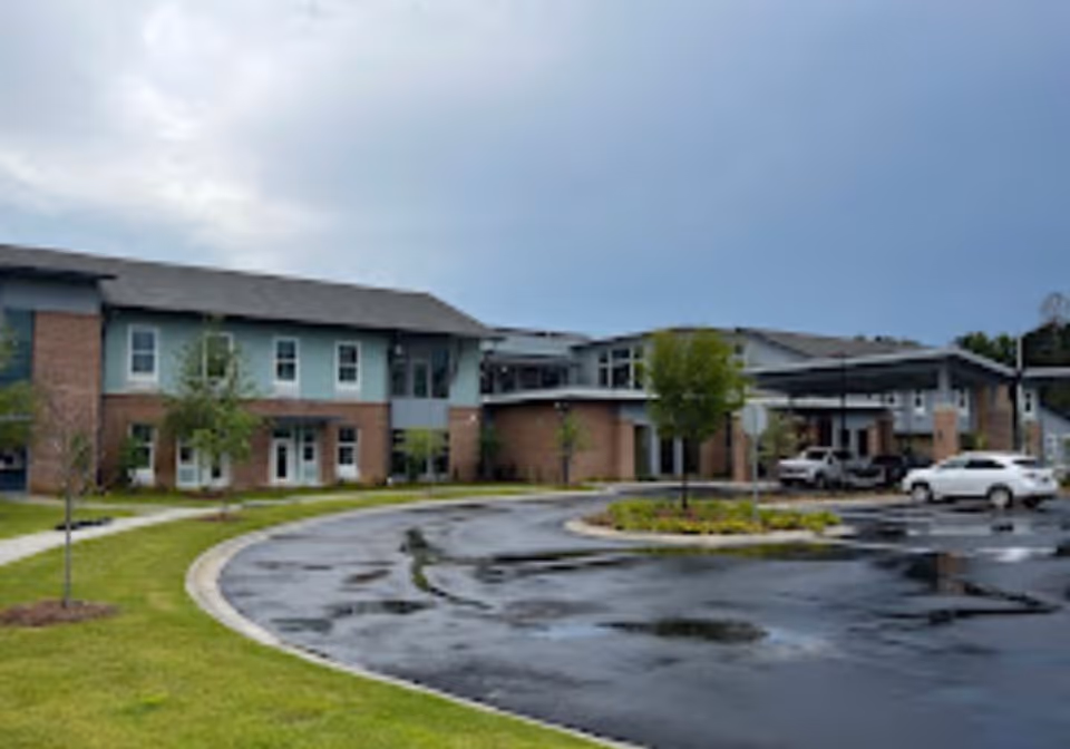 Exterior view of a two-story senior living facility building with a curved driveway and several parked cars. The building has a combination of brick and light blue siding with multiple windows and a covered entrance. The sky is cloudy and the ground appears wet from recent rain.