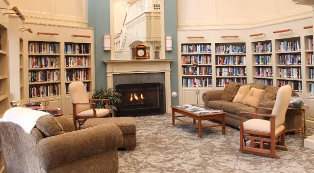 Cozy communal lounge with bookshelves lining the walls, a central fireplace, and upholstered seating around a coffee table.