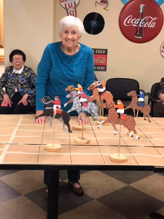 An elderly woman in a blue sweater stands behind a table with a horse racing game featuring wooden horse and jockey figures on sticks. Two other elderly women sit in chairs against the wall in the background. The room has vintage Coca-Cola signs and a vinyl record decoration on the wall.