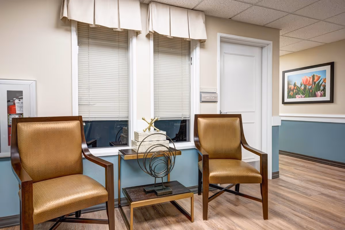 A waiting area with two brown cushioned wooden chairs placed on either side of a small table with decorative items. Behind the chairs are two windows with closed blinds and a beige valance. To the right is a white door with a sign next to it, and a framed picture of tulips hangs on the wall. The walls are painted beige and blue with white trim, and the floor is wood.