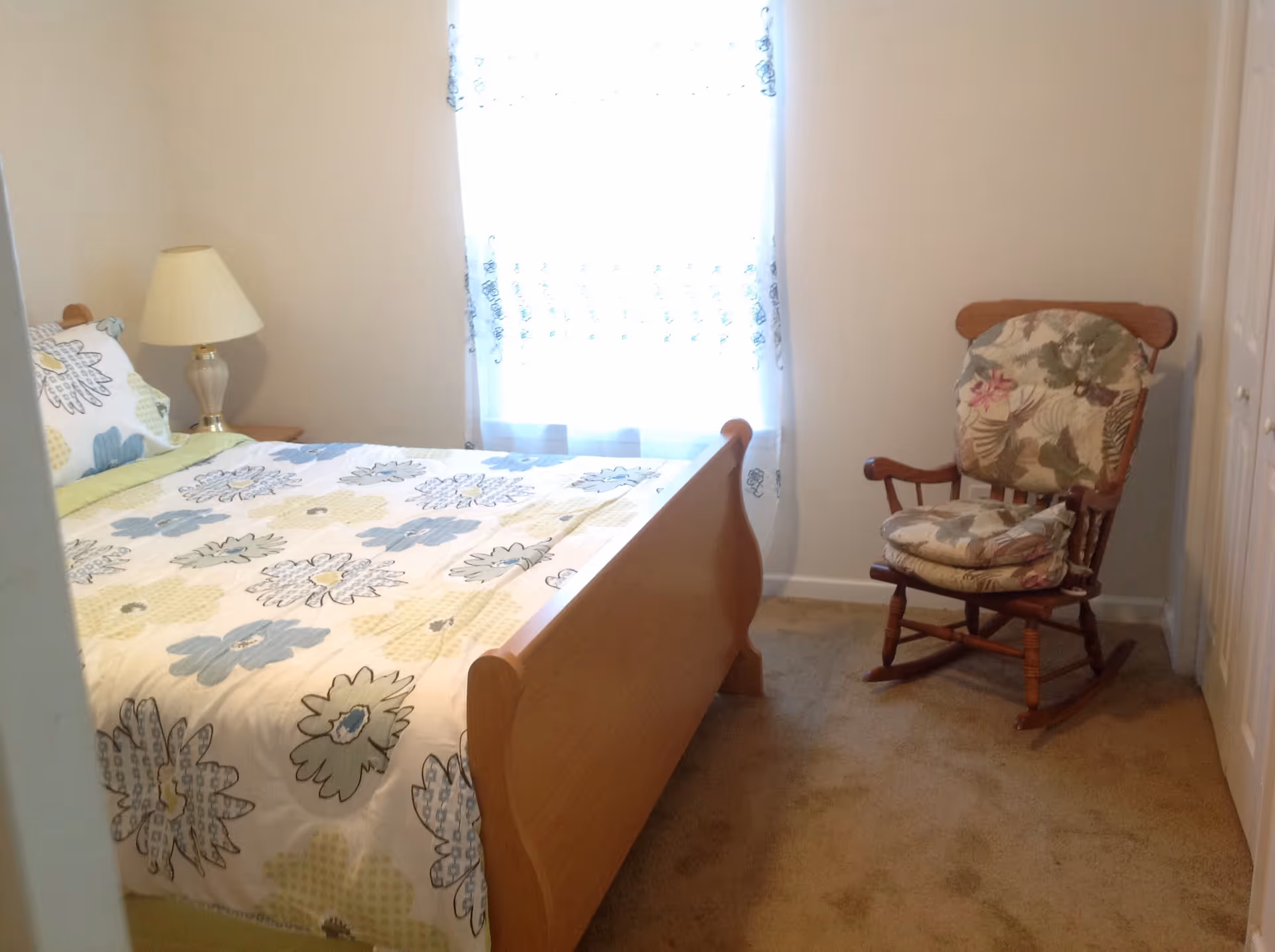 A bedroom with a wooden bed frame and floral patterned bedding in shades of blue, green, and white. There is a bedside table with a lamp on the left side of the bed. A wooden rocking chair with floral cushions is placed near a window with sheer white curtains. The room has beige carpet and light-colored walls.