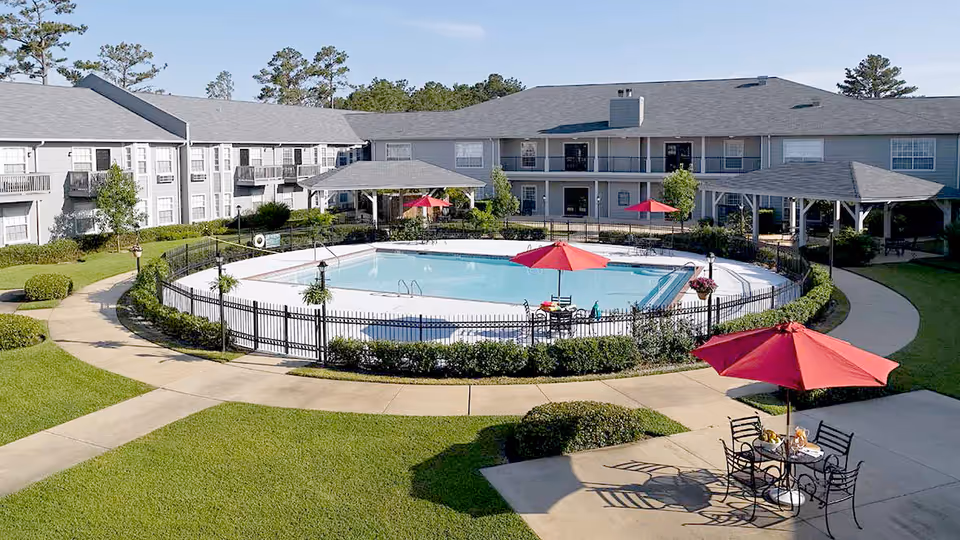 Courtyard with a central fenced swimming pool, red umbrellas and outdoor seating surrounded by a two-story residential building.