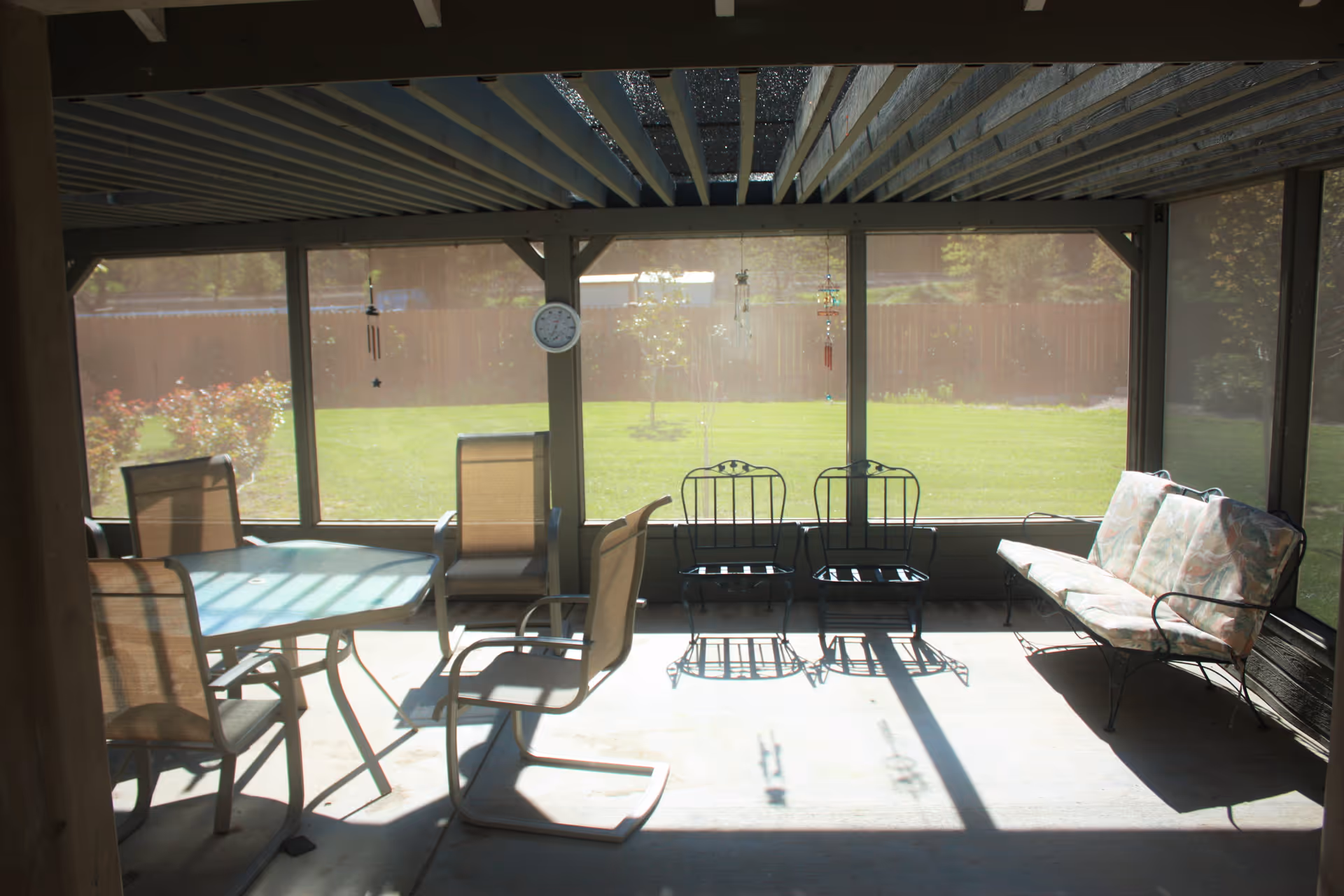 A screened-in patio area with a glass-top table and four chairs on the left side, two metal chairs in the center, and a cushioned metal bench on the right side. The patio overlooks a green lawn with a wooden fence and some bushes in the background. A clock and wind chimes hang from the patio frame.