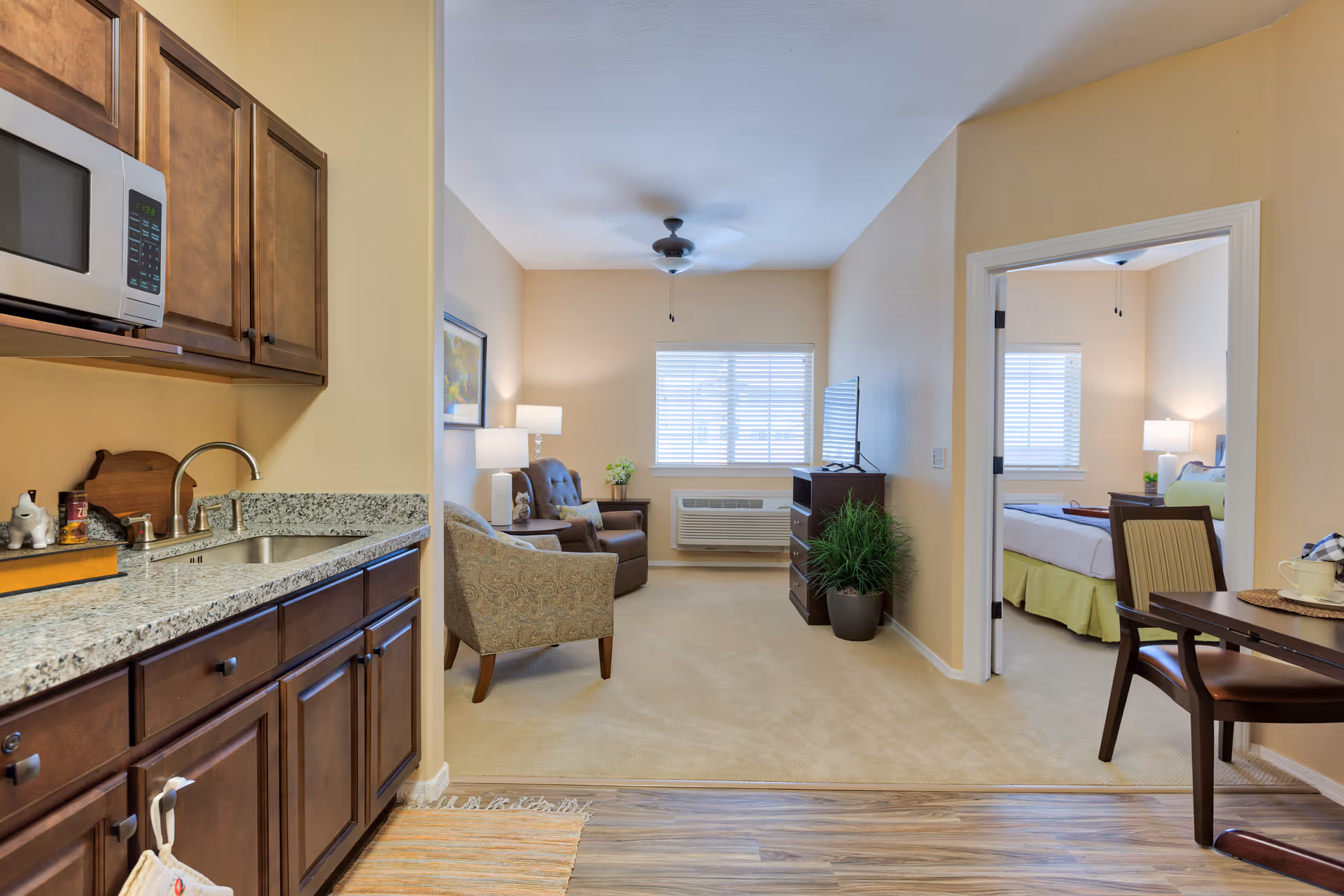 Interior view of a senior living apartment showing a small kitchen area with wooden cabinets and a microwave on the left, leading into a living room with two armchairs, a side table with a lamp, a window with blinds, and a TV on a stand. To the right, there is a dining table with chairs and a bedroom visible through an open doorway, featuring a bed, nightstand with a lamp, and a window with blinds.