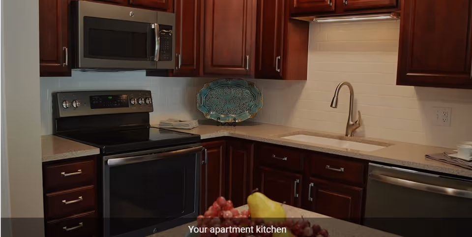Modern kitchen with dark wood cabinets, a stainless steel microwave above a black electric stove, a sink with a curved faucet, and a countertop with a decorative blue plate and a bowl of fruit including grapes and a pear.
