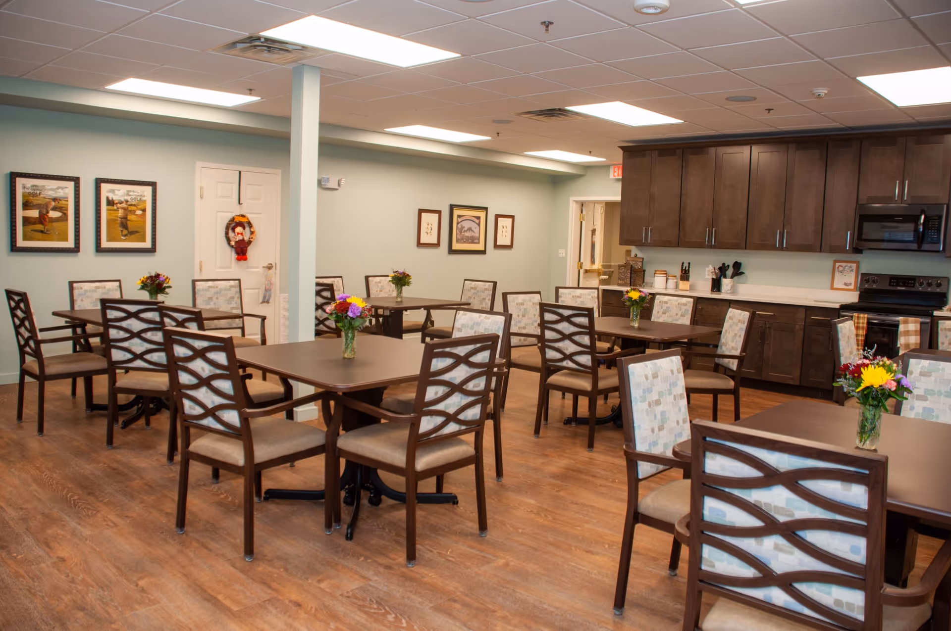 A dining room in Parkrose Estates featuring multiple square tables with wooden chairs that have patterned cushions. Each table has a small vase with colorful flowers. The room has wooden flooring, light green walls, framed pictures on the walls, and a kitchen area with dark wooden cabinets, a microwave, and an oven in the background.