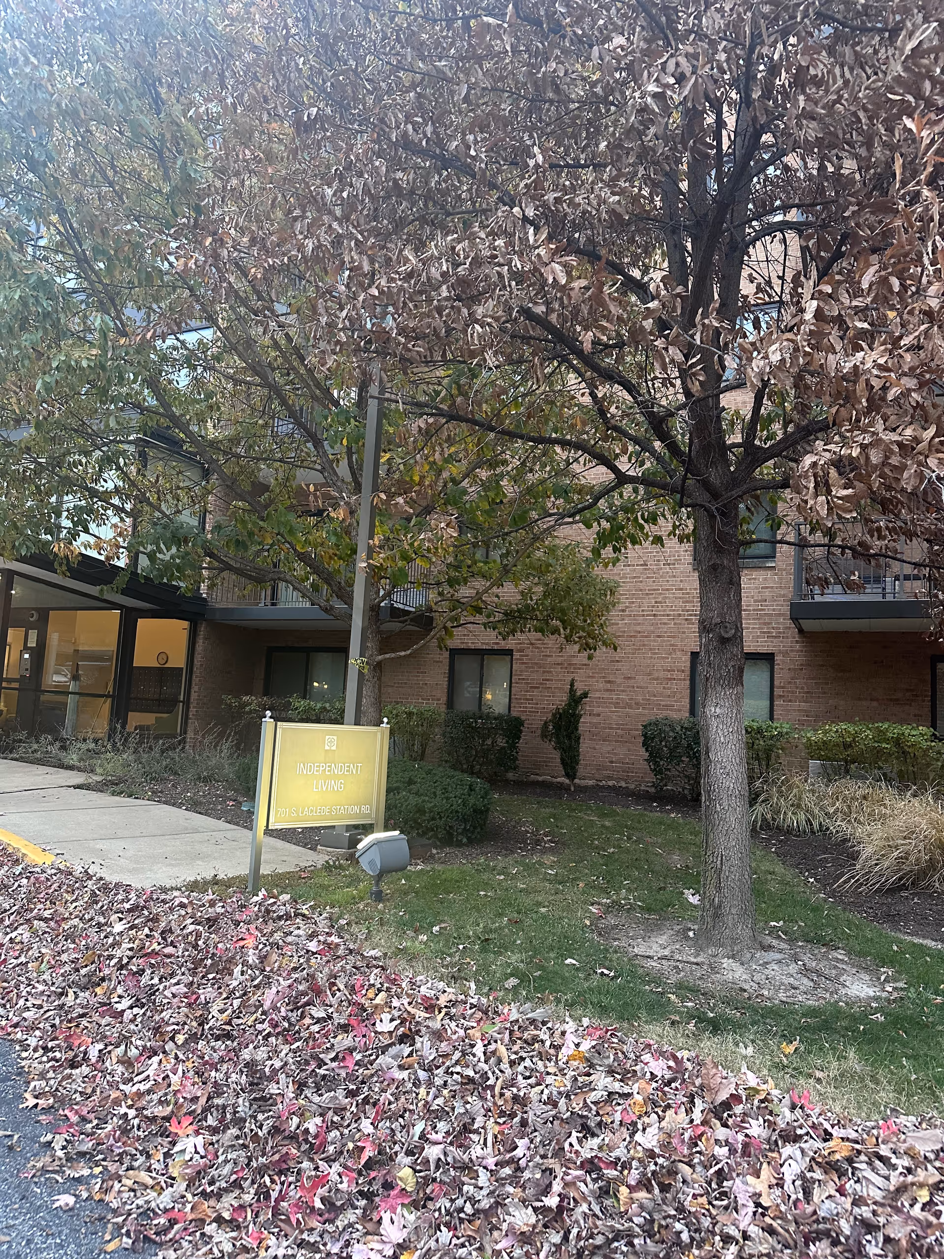 Outdoor view of the Lutheran Convalescent Home showing a brick building with windows and balconies. In front of the building, there are trees with autumn leaves, some green bushes, and a yellow sign that reads 'INDEPENDENT LIVING 701 S. LACLEDE STATION RD.'. Fallen leaves cover the ground near the sidewalk.