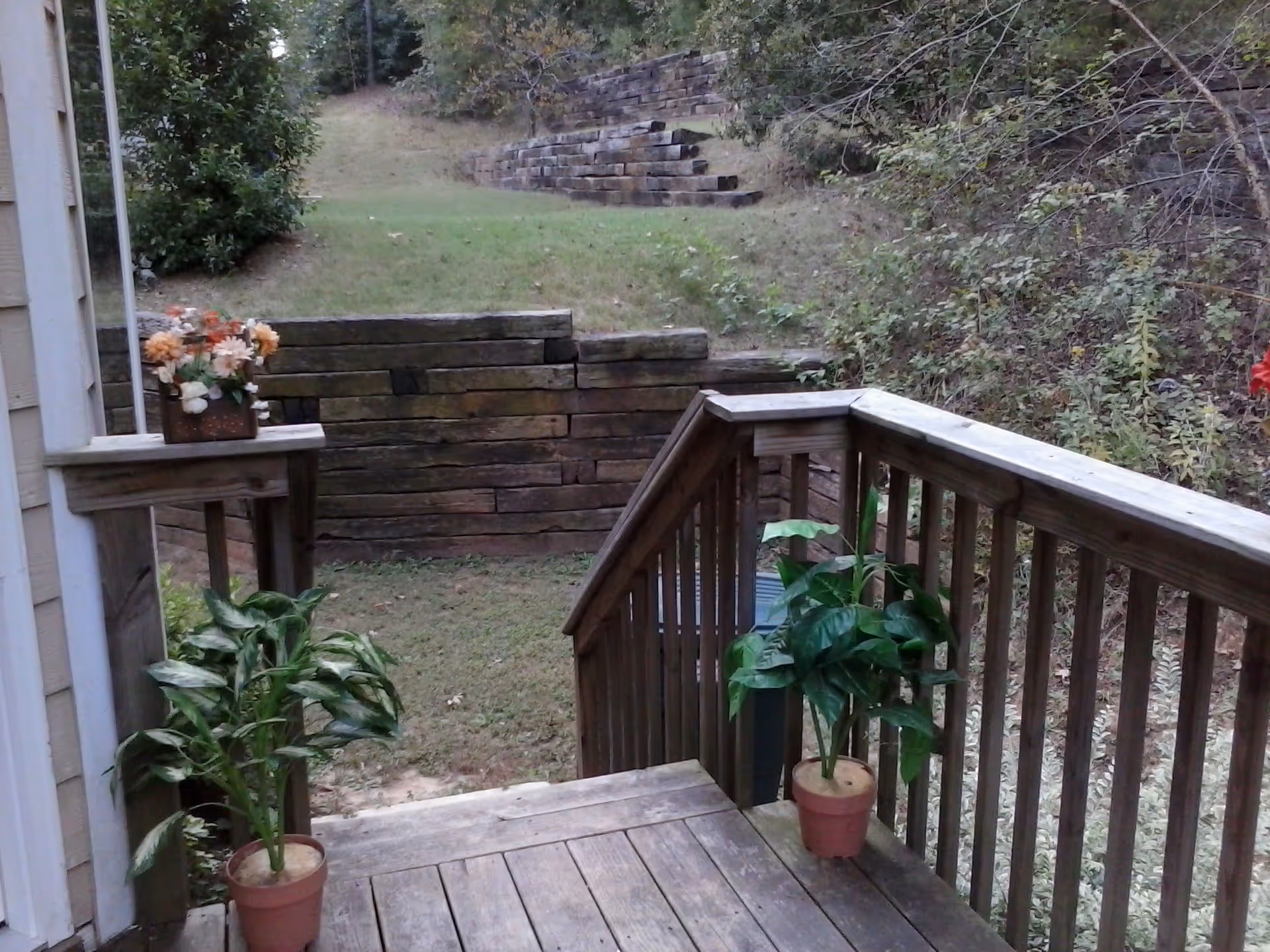 Wooden deck with two potted plants on either side of the stairs, a small wooden table with a flower arrangement on it, leading down to a grassy yard with a retaining wall and steps made of wooden beams, surrounded by trees and bushes.