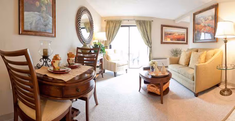 Bright furnished living room with a sofa, armchair, small round dining table, coffee table, wall art and sliding glass doors.